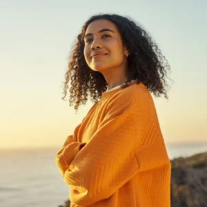 Smiling woman in an orange sweater at sunset by the coast.