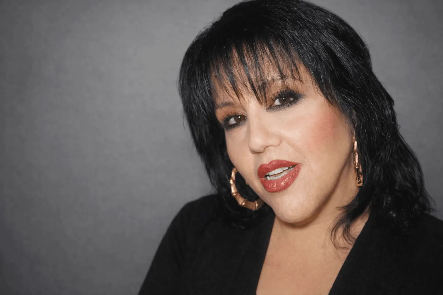 Portrait of a woman with dark hair and bold makeup against a gray background.